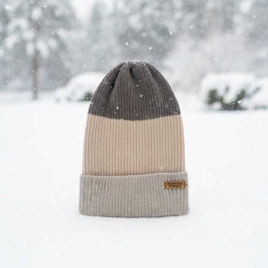 A three-tone knit beanie hat standing upright in the snow, with a dark gray top section, a beige middle section, and a light gray folded cuff. A small tan brand patch is visible on the cuff.