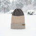 A three-tone knit beanie hat standing upright in the snow, with a dark gray top section, a beige middle section, and a light gray folded cuff. A small tan brand patch is visible on the cuff.