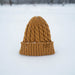 A mustard yellow cable-knit beanie hat standing upright in a snowy outdoor setting.