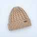 A beige cable-knit beanie hat with a folded ribbed cuff and a small gold-tone metal brand plaque on the side, shown on a plain white background.