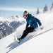 A person snowboarding down a snowy mountain slope, wearing a blue jacket, black pants, a beige beanie, and goggles, with mountains and trees in the background.