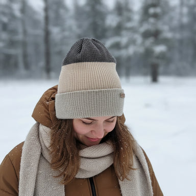 A ribbed knit beanie hat with a color-blocked design, featuring a dark brown top section, a wide beige middle section, and a light beige cuff. A small brown leather patch on the cuff reads 'HANDMADE STORIES'.