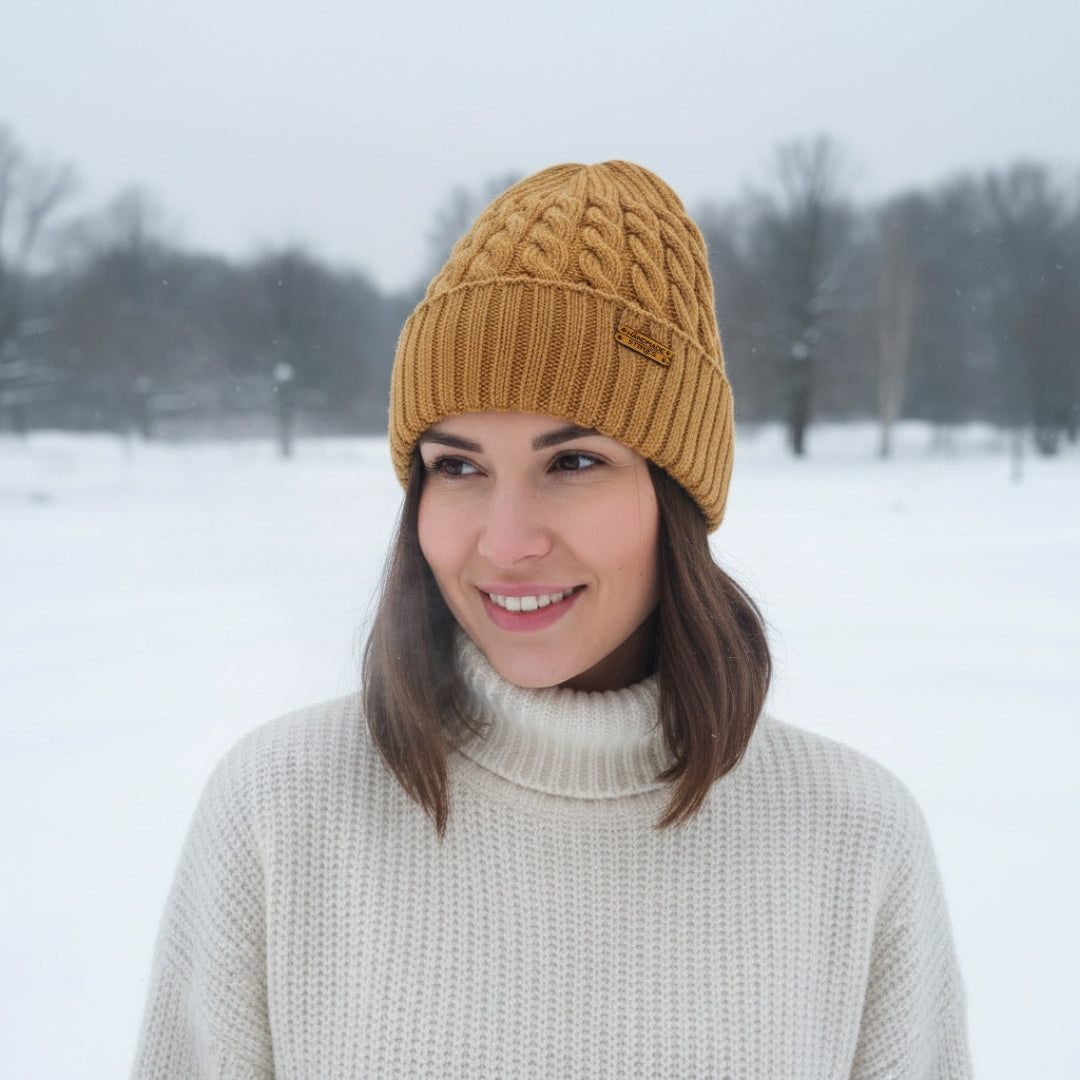 A woman wearing a mustard yellow cable-knit beanie and a cream turtleneck sweater, standing in a snowy outdoor landscape.