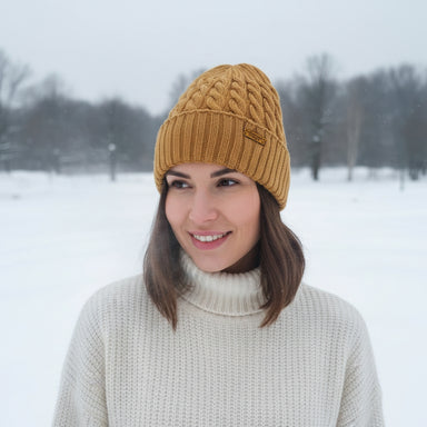 A woman wearing a mustard yellow cable-knit beanie and a cream turtleneck sweater, standing in a snowy outdoor landscape.