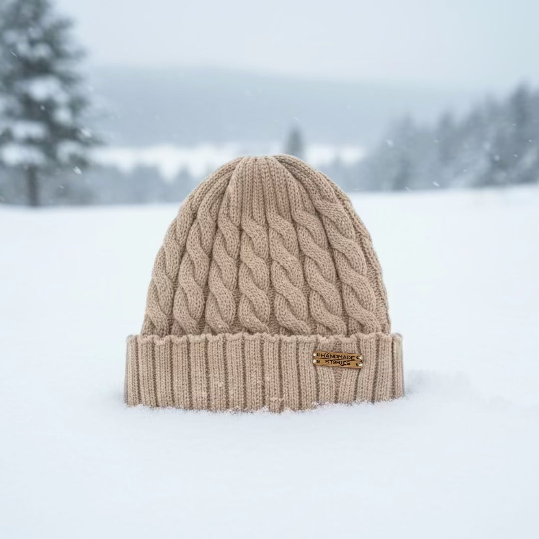 A beige cable-knit beanie hat with a folded cuff, standing in a snowy outdoor landscape with blurred trees in the background.