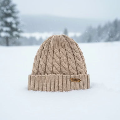 A beige cable-knit beanie hat with a folded cuff, standing in a snowy outdoor landscape with blurred trees in the background.