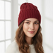 A woman wearing a chunky cable-knit beanie hat in a deep burgundy red, photographed in a bright, softly lit indoor setting.