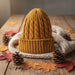 A mustard yellow cable-knit beanie hat sits on top of a cream knit scarf, styled on a rustic wooden table with autumn leaves and pinecones.